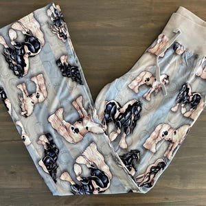 Könnte beinhalten: Hellblaue Schlafanzughose mit einem sich wiederholenden Muster von Pferden in Schwarz, Weiß und Beige. Die Hose hat einen hellgrauen Kordelzugbund. Die Pferde sind in verschiedenen Posen, mit einem Hufeisen-Design im Hintergrund.