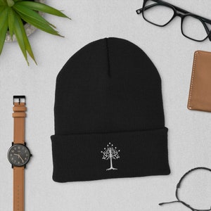 Op de afbeelding: Zwarte gebreide beanie met een wit geborduurd boomontwerp. De boom heeft een stervormige kroon en een dikke stam.