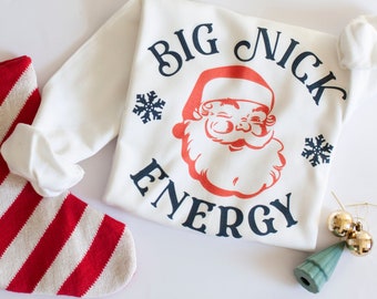 Puede incluir: Sudadera blanca con un gráfico de Papá Noel rojo y blanco y el texto "Big Nick Energy" en azul marino. La sudadera también presenta dos copos de nieve en azul marino.