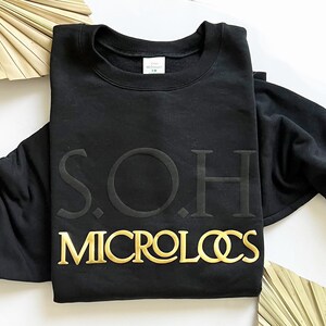 以下が含まれることがあります： 黒いスウェットシャツに、金色の文字で「S.O.H. MICROLOCS」と書かれています。