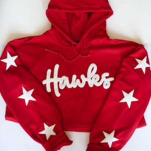 May include: A red cropped hoodie with a drawstring hood and white script text that reads "Hawks". The sleeves feature white star accents. This casual sweatshirt is perfect for showing team spirit.