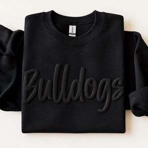 May include: A black long-sleeve crewneck sweatshirt featuring the word Bulldogs in a raised, tonal, three-dimensional puff print across the chest. The garment has a ribbed collar and a Gildan brand label visible at the inner neckline.
