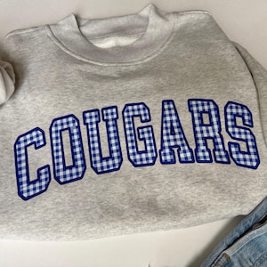 K&ouml;nnte beinhalten: Ein hellgraues Sweatshirt mit Rundhalsausschnitt und dem Wort COUGARS, das in einem blau-wei&szlig;en Vichy-Muster mit dunkelblauem Umriss gedruckt ist. Das Sweatshirt hat lange &Auml;rmel mit gerippten B&uuml;ndchen und einen gerippten Ausschnitt, kombiniert mit blauem Denim.