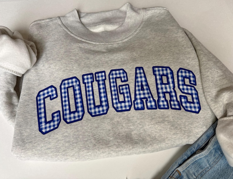 K&ouml;nnte beinhalten: Ein hellgraues Sweatshirt mit Rundhalsausschnitt und dem Wort COUGARS, das in einem blau-wei&szlig;en Vichy-Muster mit dunkelblauem Umriss gedruckt ist. Das Sweatshirt hat lange &Auml;rmel mit gerippten B&uuml;ndchen und einen gerippten Ausschnitt, kombiniert mit blauem Denim.