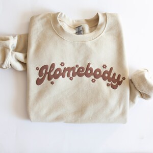 May include: A beige sweatshirt with the text "Homebody" in brown lettering with small flower accents.