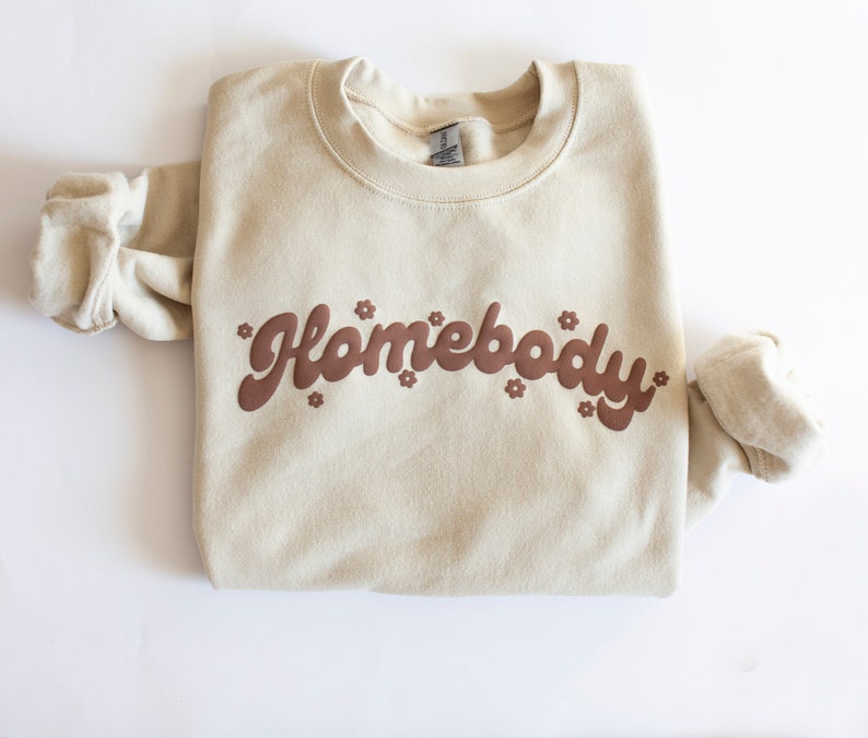 May include: A beige sweatshirt with the text "Homebody" in brown lettering with small flower accents.