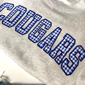 K&ouml;nnte beinhalten: Ein hellgraues Sweatshirt mit Rundhalsausschnitt und dem Wort COUGARS in gro&szlig;en Blockbuchstaben. Die Schriftz&uuml;ge haben ein blau-wei&szlig;es Vichy-Karo-Muster mit einer durchgehend dunkelblauen Umrandung, was ein markantes, college-inspiriertes Design ergibt.