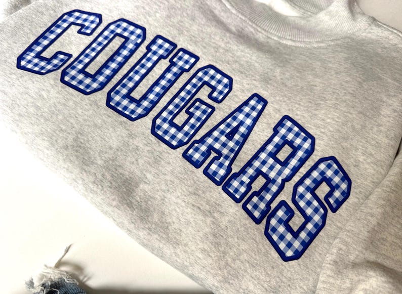 K&ouml;nnte beinhalten: Ein hellgraues Sweatshirt mit Rundhalsausschnitt und dem Wort COUGARS in gro&szlig;en Blockbuchstaben. Die Schriftz&uuml;ge haben ein blau-wei&szlig;es Vichy-Karo-Muster mit einer durchgehend dunkelblauen Umrandung, was ein markantes, college-inspiriertes Design ergibt.