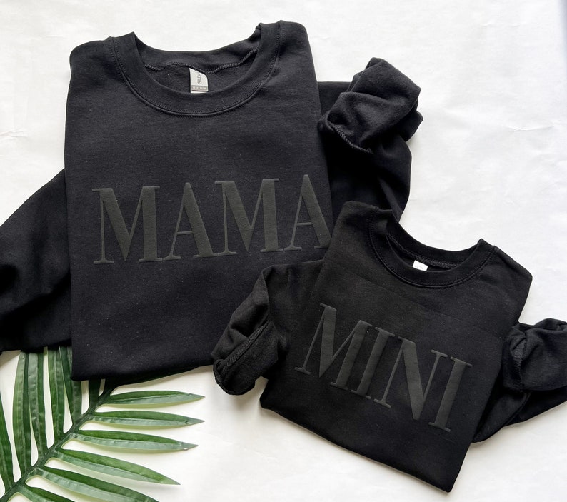 May include: Two black sweatshirts with the words "MAMA" and "MINI" printed on them. The sweatshirts are laid out on a white surface with a green plant leaf.