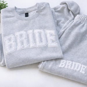 May include: A light grey sweatshirt and matching sweatpants. The word "BRIDE" is embroidered in white block letters on the front of the sweatshirt and the sweatpants. The set is made of a soft material, ideal for relaxing.
