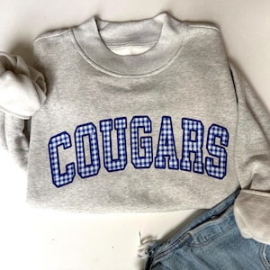 K&ouml;nnte beinhalten: Ein hellgraues Sweatshirt mit Rundhalsausschnitt und dem Wort COUGARS in einem blau-wei&szlig;en Vichy-Muster mit dunkelblauem Rand. Das Sweatshirt hat lange &Auml;rmel mit gerippten B&uuml;ndchen und einen gerippten Kragen, kombiniert mit einer blauen Jeans.