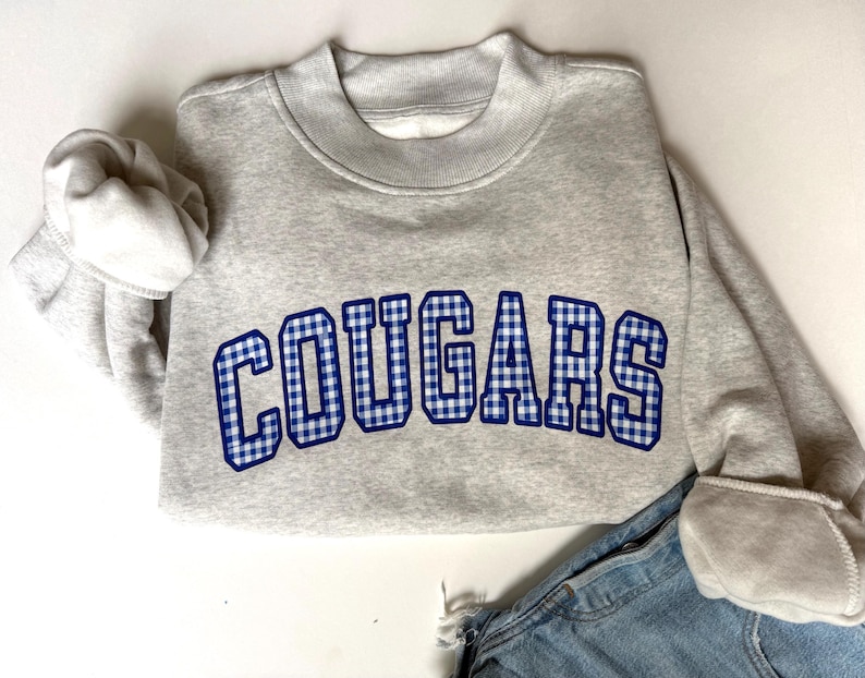 K&ouml;nnte beinhalten: Ein hellgraues Sweatshirt mit Rundhalsausschnitt und dem Wort COUGARS in einem blau-wei&szlig;en Vichy-Muster mit dunkelblauem Rand. Das Sweatshirt hat lange &Auml;rmel mit gerippten B&uuml;ndchen und einen gerippten Kragen, kombiniert mit einer blauen Jeans.