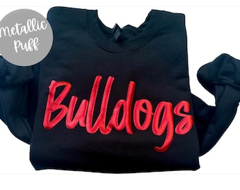 May include: Black crewneck sweatshirt with the word "Bulldogs" in red, metallic puff lettering across the chest. A gray circle with the words "Metallic Puff" is in the upper left corner.