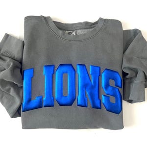May include: A gray sweatshirt with the word "LIONS" in bold, blue, raised lettering across the chest. The sweatshirt is folded neatly, with the sleeves extending to the sides. The fabric appears soft and comfortable.