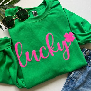 Op de afbeelding: Groen sweatshirt met een roze, cursief geschreven "lucky" en een roze klavertje vier.