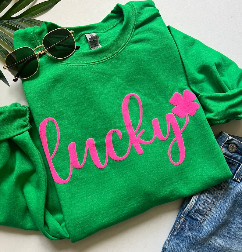 Op de afbeelding: Groen sweatshirt met een roze, cursief geschreven "lucky" en een roze klavertje vier.