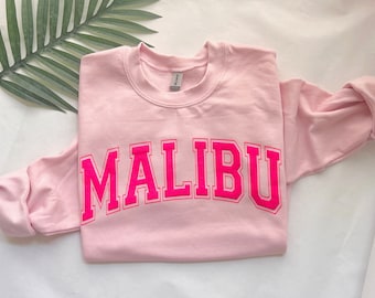 Op de afbeelding: Een lichtroze sweatshirt met het woord "MALIBU" in felroze blokletters over de borst. De sweatshirt is netjes opgevouwen, met de mouwen naar binnen. De stof ziet er zacht en comfortabel uit.