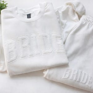 Op de afbeelding: Een witte sweatshirt en joggingbroek set. Het woord "BRIDE" is in reliëf op de voorkant van de sweatshirt en op de joggingbroek. Een kleine plant in een pot en witte stof zijn op de achtergrond.