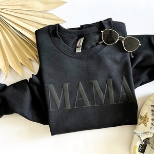 May include: Black sweatshirt with the word "MAMA" in large, bold, black letters. The sweatshirt is laid flat on a white surface with a pair of sunglasses and a white sneaker with a gold star on the side.