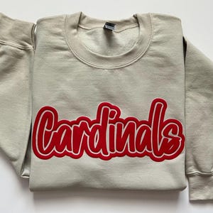 Puede incluir: Una sudadera gris claro con la palabra "Cardinals" en rojo, contorneada en blanco, en el pecho. La sudadera tiene cuello redondo y mangas largas. La tela parece suave y cómoda.