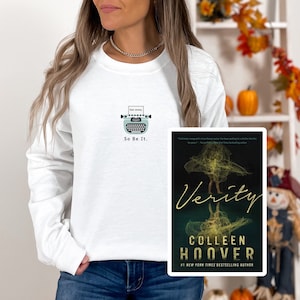 May include: White long-sleeve sweatshirt with a teal typewriter graphic and the text "Dear Jeremy. So Be It." Next to the sweatshirt is a book cover for "Verity" by Colleen Hoover.