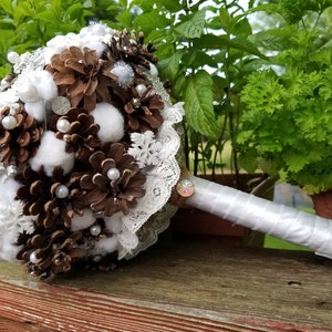 May include: A white and brown wedding bouquet made of pine cones, cotton balls, lace, and pearls. The bouquet has a white satin ribbon handle.