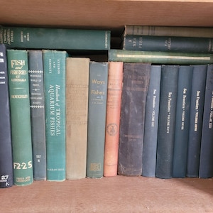 May include: A collection of hardcover books arranged on a wooden shelf. The spines feature various shades of green, blue, tan, and brown, with titles including Wildlife in Danger, Fish and Fisheries of Australia, Handbook of Tropical Aquarium Fishes, and Sea Frontiers.