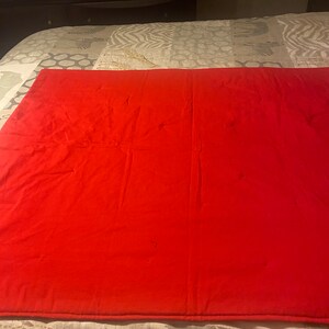 Puede incluir: Una colcha roja s&oacute;lida con un sutil patr&oacute;n acolchado. La colcha est&aacute; doblada por la mitad y puesta sobre una cama con una colcha estampada.