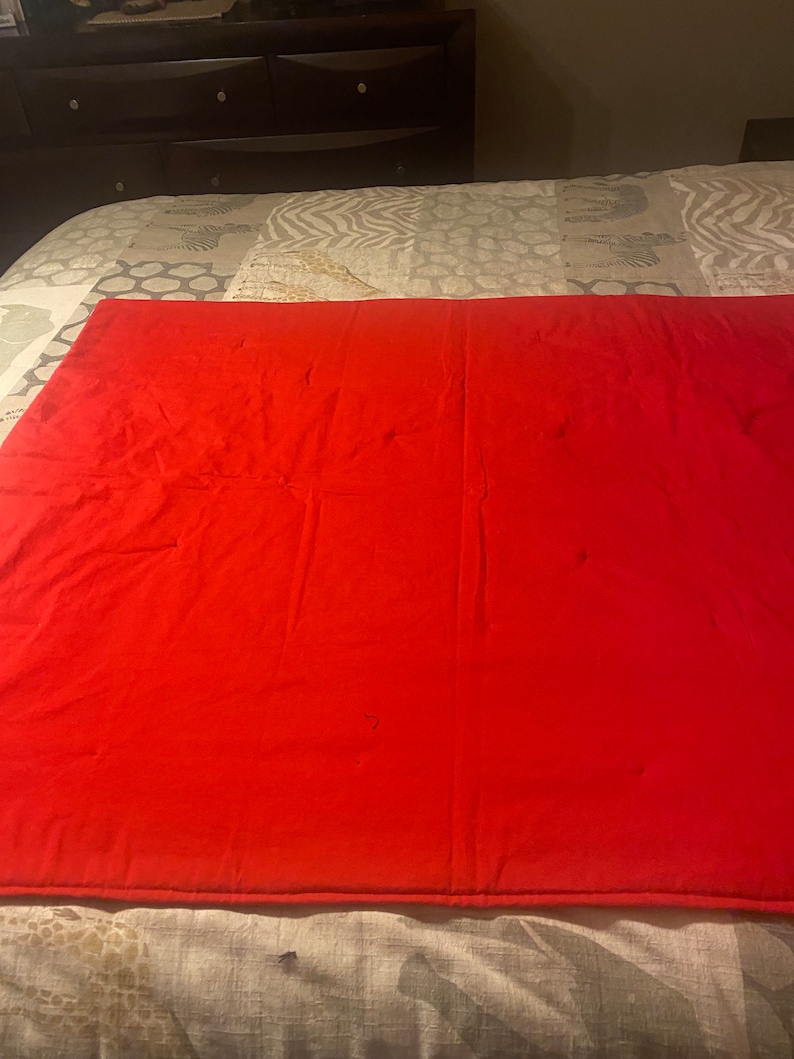 Puede incluir: Una colcha roja s&oacute;lida con un sutil patr&oacute;n acolchado. La colcha est&aacute; doblada por la mitad y puesta sobre una cama con una colcha estampada.