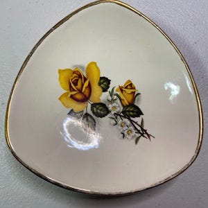 May include: A white triangular ceramic dish with a gold rim. The dish features a yellow rose with a red centre and a smaller yellow rose with a green stem and white flowers.