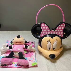 Puede incluir: Un juguete de peluche Minnie Mouse rosa y negro acostado en una manta rosa y verde con un cubo Minnie Mouse rosa y negro con un asa rosa.