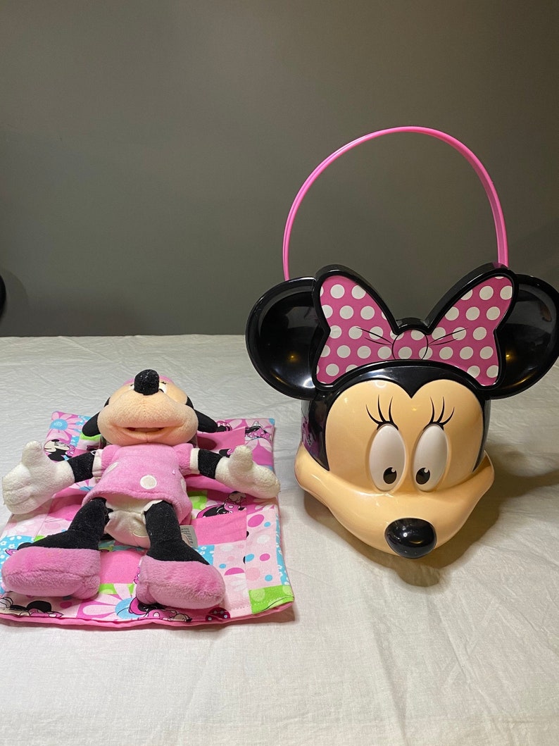 Puede incluir: Un juguete de peluche Minnie Mouse rosa y negro acostado en una manta rosa y verde con un cubo Minnie Mouse rosa y negro con un asa rosa.