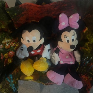 Puede incluir: Un conjunto de juguetes de peluche de Mickey Mouse y Minnie Mouse. Mickey lleva una camisa roja con lunares blancos y zapatos amarillos. Minnie lleva un vestido rosa con lunares blancos y un lazo rosa. Los juguetes est&aacute;n sentados sobre un fondo de tela colorido.