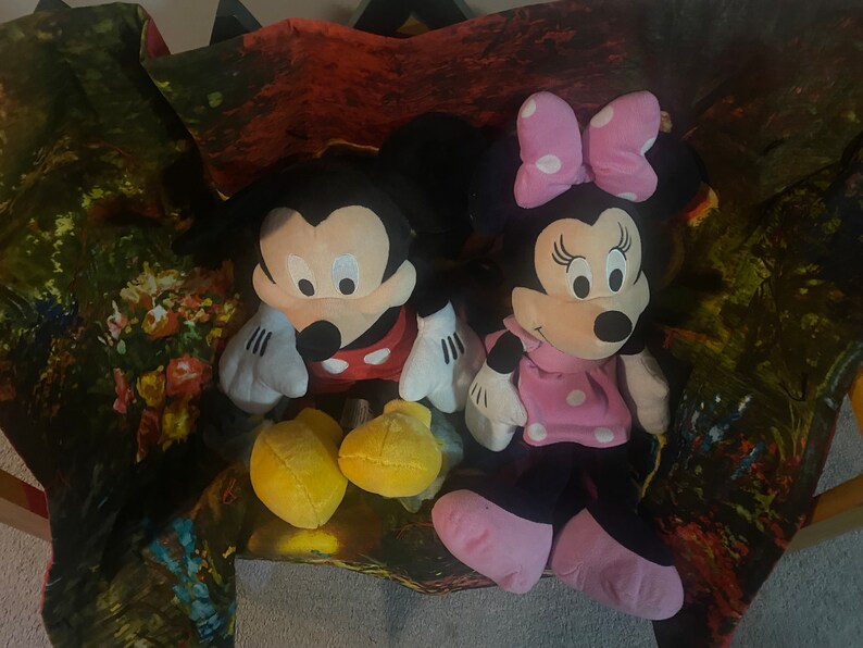 Puede incluir: Un conjunto de juguetes de peluche de Mickey Mouse y Minnie Mouse. Mickey lleva una camisa roja con lunares blancos y zapatos amarillos. Minnie lleva un vestido rosa con lunares blancos y un lazo rosa. Los juguetes est&aacute;n sentados sobre un fondo de tela colorido.