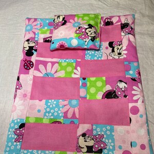Puede incluir: Una cama de mu&ntilde;eca de retazos rosa y verde con una almohada a juego con un dise&ntilde;o de Minnie Mouse. La cama est&aacute; hecha de tela con una variedad de patrones, incluyendo lunares, flores y caras de Minnie Mouse.