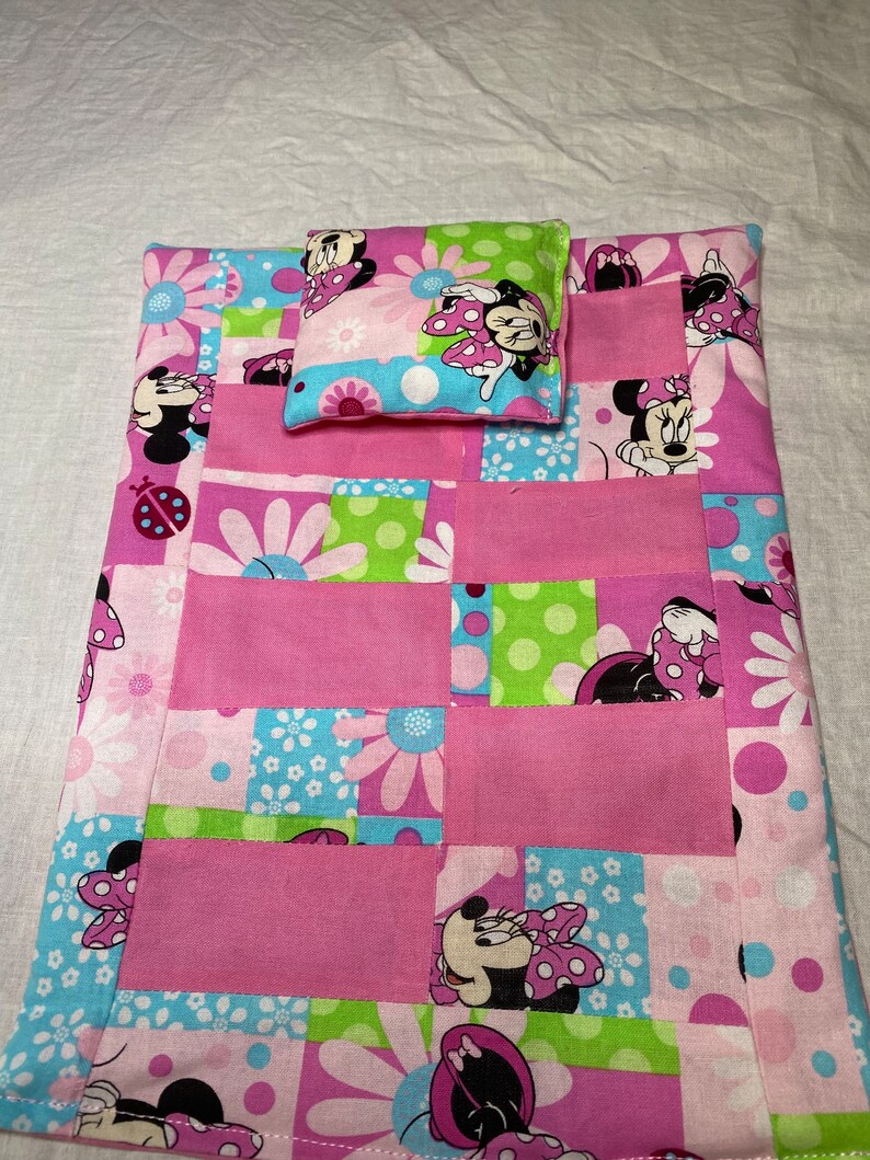Puede incluir: Una cama de mu&ntilde;eca de retazos rosa y verde con una almohada a juego con un dise&ntilde;o de Minnie Mouse. La cama est&aacute; hecha de tela con una variedad de patrones, incluyendo lunares, flores y caras de Minnie Mouse.