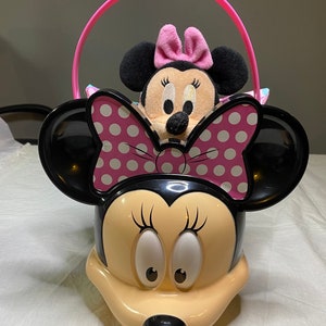 Puede incluir: Un cubo de pl&aacute;stico rosa y negro con forma de personaje de Minnie Mouse con un asa rosa. Un juguete de peluche Minnie Mouse m&aacute;s peque&ntilde;o est&aacute; sentado dentro del cubo.