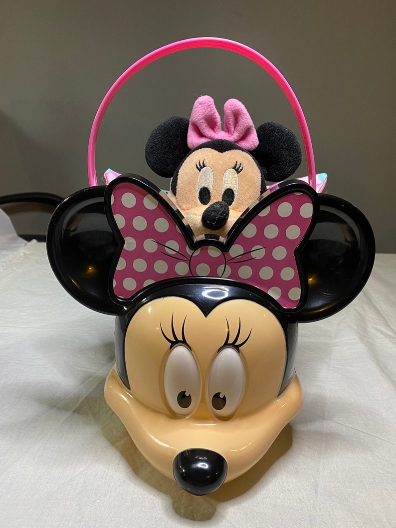 Puede incluir: Un cubo de pl&aacute;stico rosa y negro con forma de personaje de Minnie Mouse con un asa rosa. Un juguete de peluche Minnie Mouse m&aacute;s peque&ntilde;o est&aacute; sentado dentro del cubo.