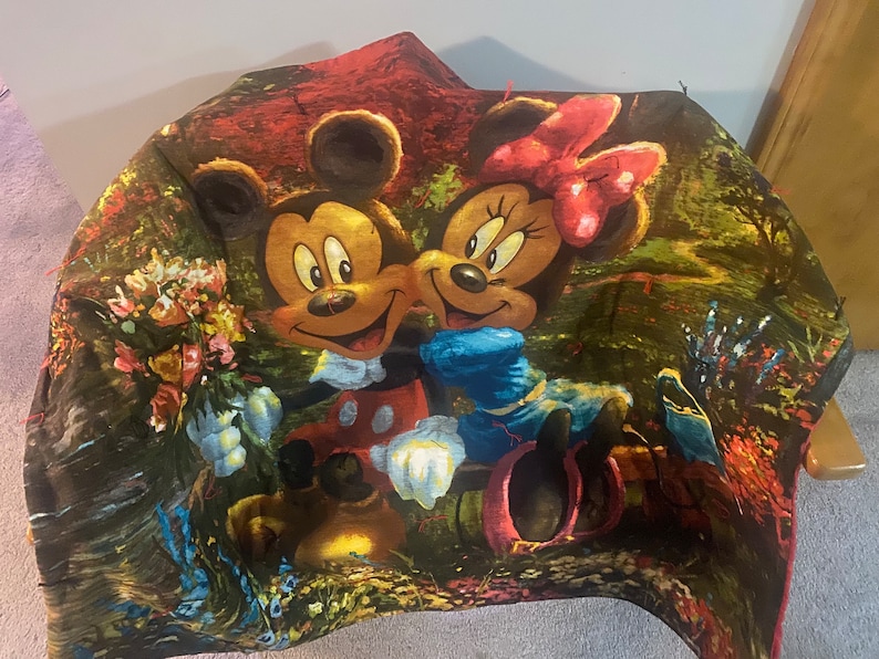 Puede incluir: Una manta de felpa colorida con Mickey Mouse y Minnie Mouse en un entorno de bosque de ensue&ntilde;o. La manta est&aacute; hecha de tela suave y tiene un esquema de color verde, rojo y amarillo vibrante.