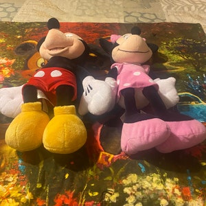 Puede incluir: Un conjunto de juguetes de peluche de Mickey Mouse y Minnie Mouse. Mickey lleva pantalones cortos rojos con lunares blancos y zapatos amarillos. Minnie lleva un vestido rosa con lunares blancos y zapatos negros.