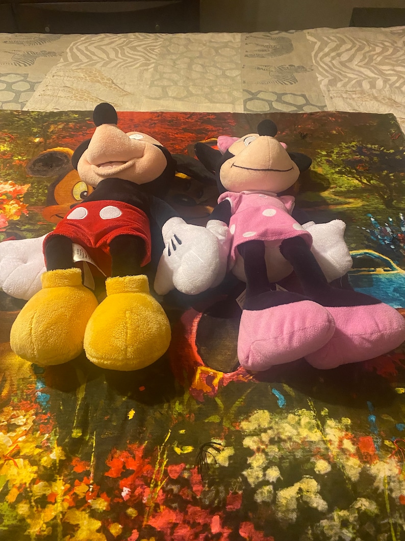 Puede incluir: Un conjunto de juguetes de peluche de Mickey Mouse y Minnie Mouse. Mickey lleva pantalones cortos rojos con lunares blancos y zapatos amarillos. Minnie lleva un vestido rosa con lunares blancos y zapatos negros.