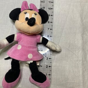 Puede incluir: Un juguete de peluche rosa y negro de Minnie Mouse. Lleva un vestido rosa con lunares blancos y un lazo negro. Tiene una nariz negra y grandes ojos negros.