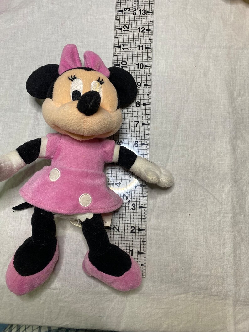 Puede incluir: Un juguete de peluche rosa y negro de Minnie Mouse. Lleva un vestido rosa con lunares blancos y un lazo negro. Tiene una nariz negra y grandes ojos negros.