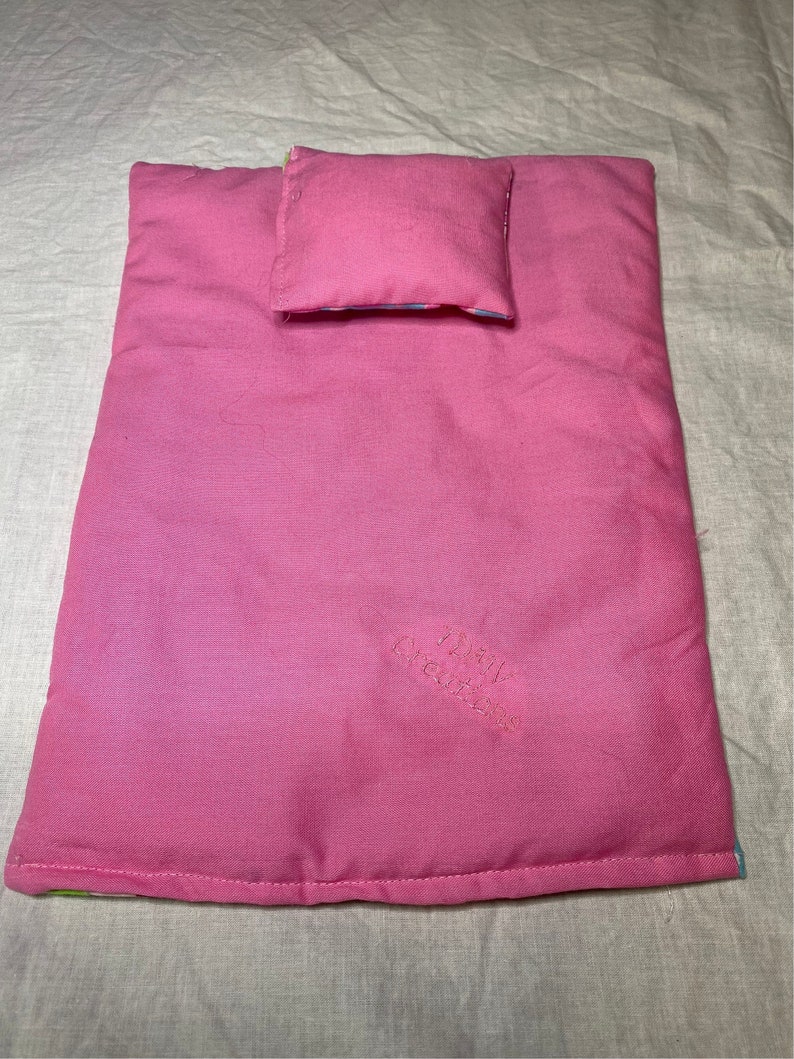 Puede incluir: Una cama de mu&ntilde;eca rosa con una almohada rosa a juego. La cama est&aacute; bordada con el texto "Pink Creations".