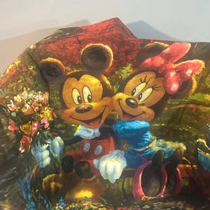 Puede incluir: Una tela colorida con Mickey Mouse y Minnie Mouse en un entorno de jard&iacute;n. La tela tiene un fondo verde, marr&oacute;n y rojo con flores y plantas.