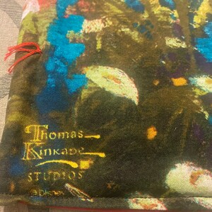 Puede incluir: Primer plano de una tela verde oscura con un dise&ntilde;o abstracto azul y amarillo. La tela tiene una etiqueta bordada en oro que dice "Thomas Kinkade Studios".