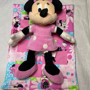 Puede incluir: Un juguete de peluche rosa y negro de Minnie Mouse est&aacute; acostado sobre una colcha de retazos rosa y verde. La colcha tiene un patr&oacute;n floral con im&aacute;genes de Minnie Mouse.