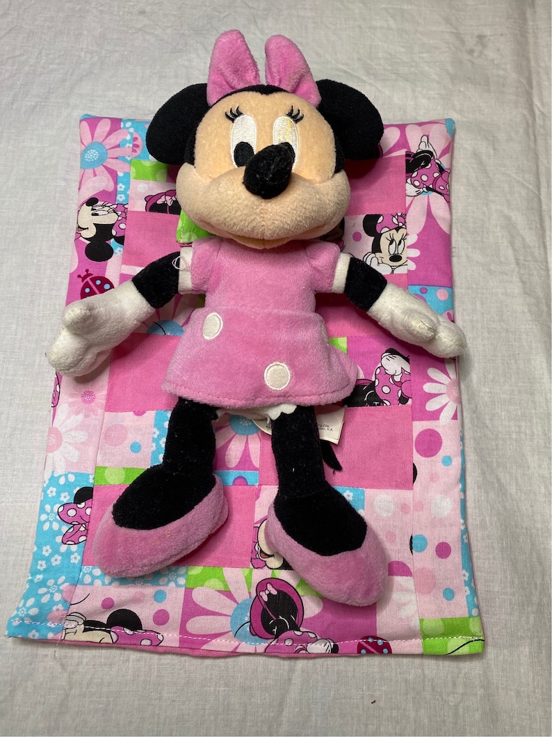 Puede incluir: Un juguete de peluche rosa y negro de Minnie Mouse est&aacute; acostado sobre una colcha de retazos rosa y verde. La colcha tiene un patr&oacute;n floral con im&aacute;genes de Minnie Mouse.