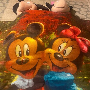 Puede incluir: Una manta roja, verde y marr&oacute;n con una imagen impresa de Mickey Mouse y Minnie Mouse. La manta presenta un primer plano de sus caras y un lazo azul en Minnie Mouse.