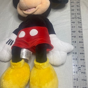 Puede incluir: Un juguete de peluche de Mickey Mouse con pelaje negro, pantalones cortos rojos con lunares blancos y zapatos amarillos. El juguete mide aproximadamente 50 cm de alto.